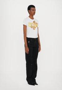 Balta marškinėliai su auksiniu "Versace Jeans Couture" logotipu, derinami su juodais tekstūriniais džinsais, turinčiais šoninį užtrauktuką ir dirželio detalę.