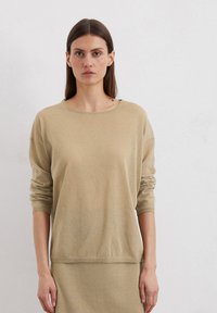 Haut beige à manches longues en tissu doux, avec une coupe décontractée. Il présente un col rond et des épaules tombantes, avec des poignets et un ourlet côtelés.