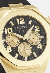 Reloj analógico en tono dorado con esfera negra texturizada, grandes marcadores de hora dorados, tres subesferas y una resistente correa de goma negra. Caja redonda.