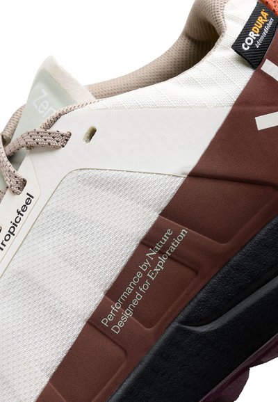 Gros plan sur une sneaker blanche et marron avec des lacets beige, comportant le texte "Performance par Nature Conçu pour l'Exploration" et une étiquette en tissu Cordura.