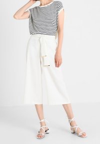 Pruhovaný krátký rukáv top v bílé a námořnické modři, kombinovaný s bílými culottes s vysokým pasem a zavázaným opaskem. Bílá sandály na tenké pásky dokončují vzhled.