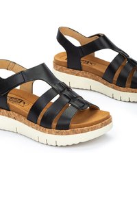 Sandalias de cuero negro con múltiples correas, plantilla de corcho y suela de goma blanca. Presentan una textura suave y un diseño de talón abierto.