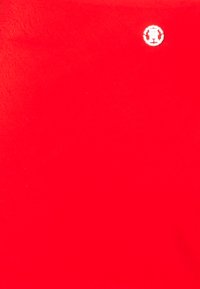 Vörös textúrált anyag, amelyen a jobb felső sarokban egy kis ezüst embléma található, sima és egyenletes felülettel.