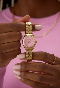 Reloj Timex de tono dorado con una esfera rosa, bisel estriado y pulsera enlazada sostenido entre dos manos. Las manos presentan una ligera manicura.