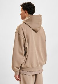 Sudadera con capucha en tejido marrón claro, con un corte relajado, puños de canalé y cinturilla, con una textura suave y un diseño minimalista.