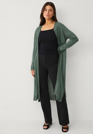 Lang, åben-front cardigan i blødt grønt stof med ribbet tekstur, parret med en sort tanktop og sorte flagrende bukser.