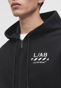 Gros plan d'une personne portant un sweat à capuche noir zippé, avec un texte blanc et un motif sur la poitrine indiquant I./AB c/o Off-White.