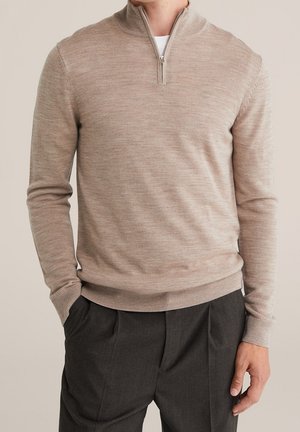 Man draagt een beige lange mouwen sweater met rits en donkergrijze geplooide broek, met één hand in de zak, tegen een neutrale achtergrond.