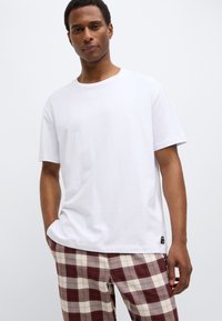 Weißes Baumwoll-T-Shirt mit Rundhalsausschnitt, kurzen Ärmeln und legerer Passform, kombiniert mit rot-beigen karierte Pyjamahosen.