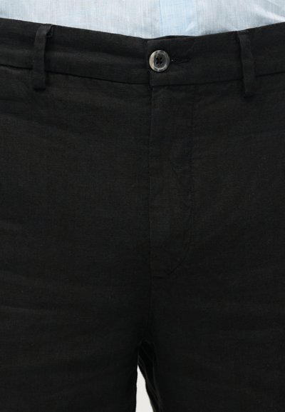 Κοντινή λήψη μαύρου παντελονιού με κουμπί και θηλιές για ζώνη, φορεμένο με ανοιχτόχρωμο πουκάμισο μέσα.