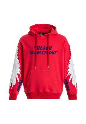 Joven con sudadera roja con diseños de alas blancas en las mangas, pantalones cortos beige con cadena, calcetines blancos y zapatos beige texturizados, de pie recto.