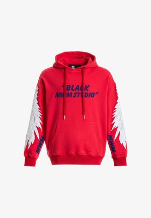 Joven con sudadera roja con diseños de alas blancas en las mangas, pantalones cortos beige con cadena, calcetines blancos y zapatos beige texturizados, de pie recto.