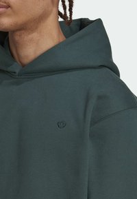 Närbild på person som bär en mörkgrön hoodie med en liten broderad treklöverlogotyp på bröstet, visar nacke och del av ansikte.
