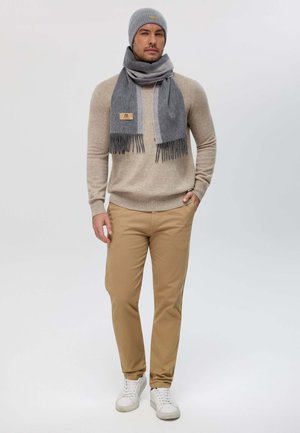 Maglione beige chiaro, berretto grigio a maglia, sciarpa coordinata con frange, pantaloni chinos beige e sneakers bianche. La sciarpa ha un'etichetta.