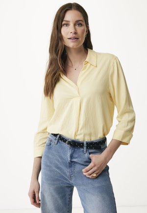Camicia gialla con colletto e maniche lunghe, abbinata a jeans azzurri chiari e una cintura nera con pois bianchi.