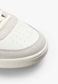 Vârf de pantof sport din piele albă cu accente din suede gri, talpă din cauciuc texturat bej și perforații mici pe partea superioară pentru respirabilitate.