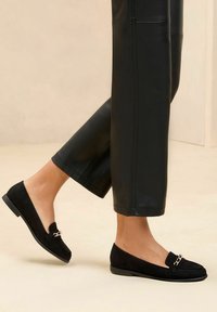 Schwarze Wildleder-Lederhalbschuhe mit flacher Sohle und dekorativem Kettenakzent. Getragen mit cropped schwarzen Lederhosen. Glatte Textur und einfaches Design.