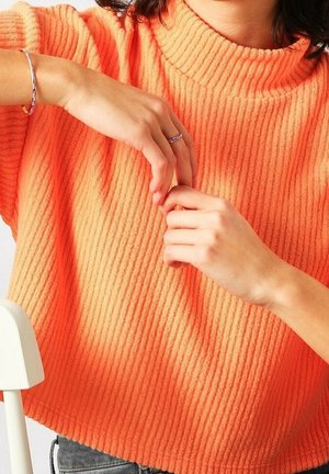 Personne portant un pull orange côtelé, montrant des mains avec une bague et un bracelet en argent, ajustant les doigts contre le dossier blanc d'une chaise.