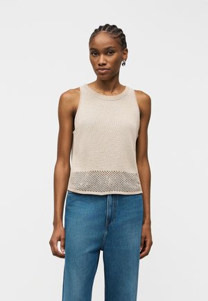 Žena s vrkočmi, oblečená v béžovom bezrukávovom pletenom topu s priesvitným lemom a modrých džínsach s vysokým pásom, stojaca na jednoduchom pozadí.