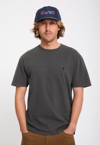 Jeune homme aux cheveux mi-longs, portant un t-shirt gris foncé, un pantalon en velours côtelé marron et une casquette bleu foncé avec une broderie colorée.