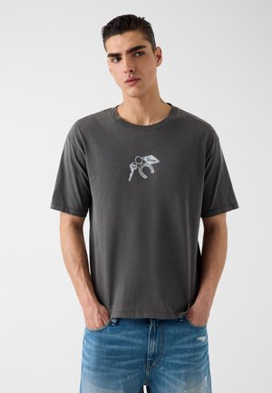 Giovane uomo che indossa una maglietta nera sbiadita con una piccola grafica di chiavi e un ferro di cavallo, abbinata a jeans di denim blu, con le mani in tasca.