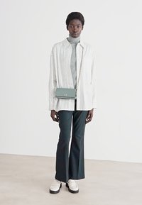 White and gray striped oversized shirt, gray turtleneck, green crossbody bag, black flared pants, and beige loafers. Minimalist styling.