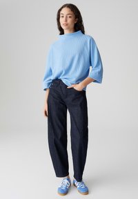 Hellblaues, geripptes Strickoberteil mit hohem Kragen; kombiniert mit dunkelblauen, weit geschnittenen Jeans. Die Schuhe sind blaue Sneaker mit weißen Schnürsenkeln.