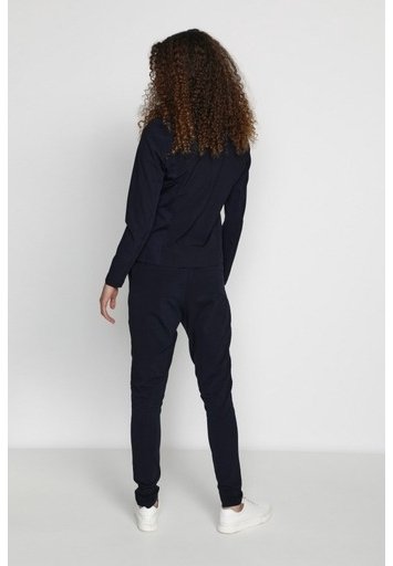 Personne aux longs cheveux bouclés, debout de dos, portant une veste noire cintrée, un pantalon noir et des baskets blanches sur un fond uni.