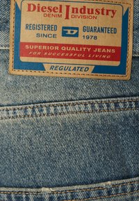 Jeans din denim cu o etichetă din piele maro care prezintă text roșu și albastru. Eticheta are inscripția "Diesel Industry" și include detalii despre marcă și calitate.