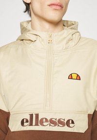 Personne portant un sweat à capuche beige et marron avec fermeture éclair devant et logo ellesse sur la poitrine et le rabat de poche.