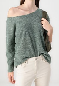 Pull vert texturé à épaules dénudées, à coupe ample, avec des manches longues et un motif pied-de-poule. Associé à un pantalon de couleur claire.