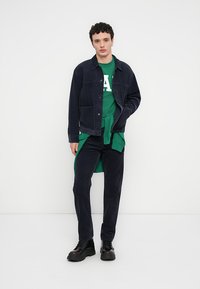 Μπλε σκούρο τζάκετ κορντόνι πάνω από πράσινο φούτερ με λευκό κείμενο, σε συνδυασμό με μαύρο παντελόνι και χοντρά μαύρα μποτάκια, στέκεται μπροστά σε απλό φόντο.