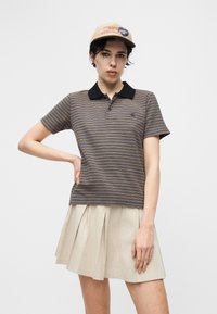 Γυναίκα που φοράει μπεζ καπέλο, ριγέ πόλο πουκαμίσα με μαύρο γιακά και μπεζ πλισέ φούστα, στέκεται με το ένα χέρι στη μέση μπροστά σε απλό φόντο.