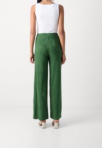 Zelené široké kalhoty s texturovaným vzorem listů palmy; doplněné bílým přiléhavým tílkem a světle barevnými sandály s blokovým podpatkem.