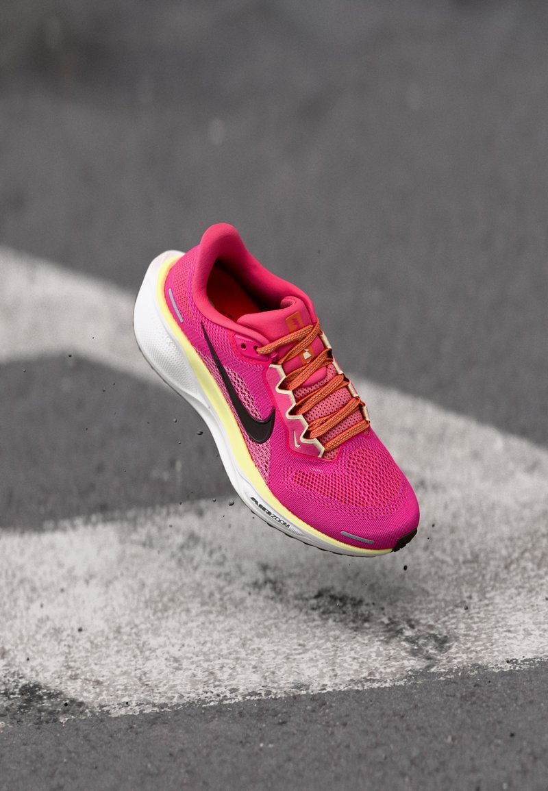 Chaussure de sport rose vif avec des lacets orange et un logo noir, en l'air au-dessus d’un bitume gris avec des marquages routiers blancs.