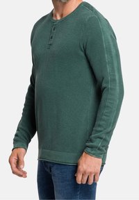 Zelená dlouhá rukávová triko henley s texturovaným pleteným vzorem, s trojicí knoflíků a žebrovaným lemem a manžetami; nošeno přes džíny.