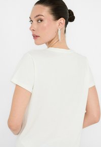 Camiseta blanca de manga corta con un cuello redondo. La tela parece suave, y hay unos pendientes brillantes y colgantes visibles en el fondo.