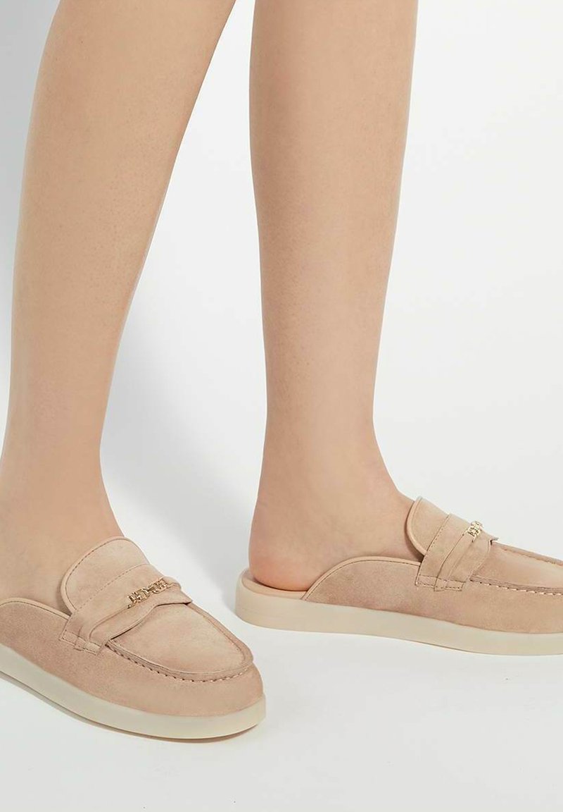 Beige suède instappers met een ronde neus, een laag profiel rubberen zool en een decoratieve band met een metalen accent.