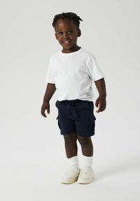 T-shirt blanc à manches courtes, short cargo bleu marine avec poches latérales, chaussettes blanches mi-mollet et chaussures slip-on beiges. Debout avec un sourire.