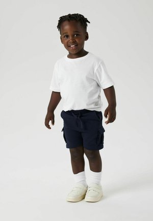 Camiseta blanca de manga corta, pantalones cortos de carga marinos con bolsillos laterales, calcetines blancos hasta el tobillo y zapatos tipo slip-on beige. De pie con una sonrisa.