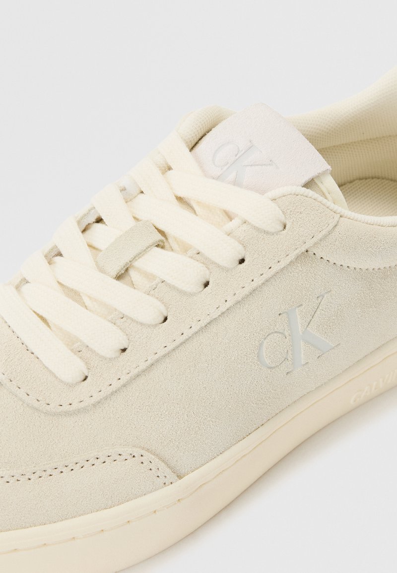 Beżowy sneaker zamszowy z zaokrąglonym noskiem, płaskimi sznurowadłami, wyściełanym kołnierzem i subtelnym akcentem logo. Podeszwa jest gładka, podkreślając czyste linie i prosty design.