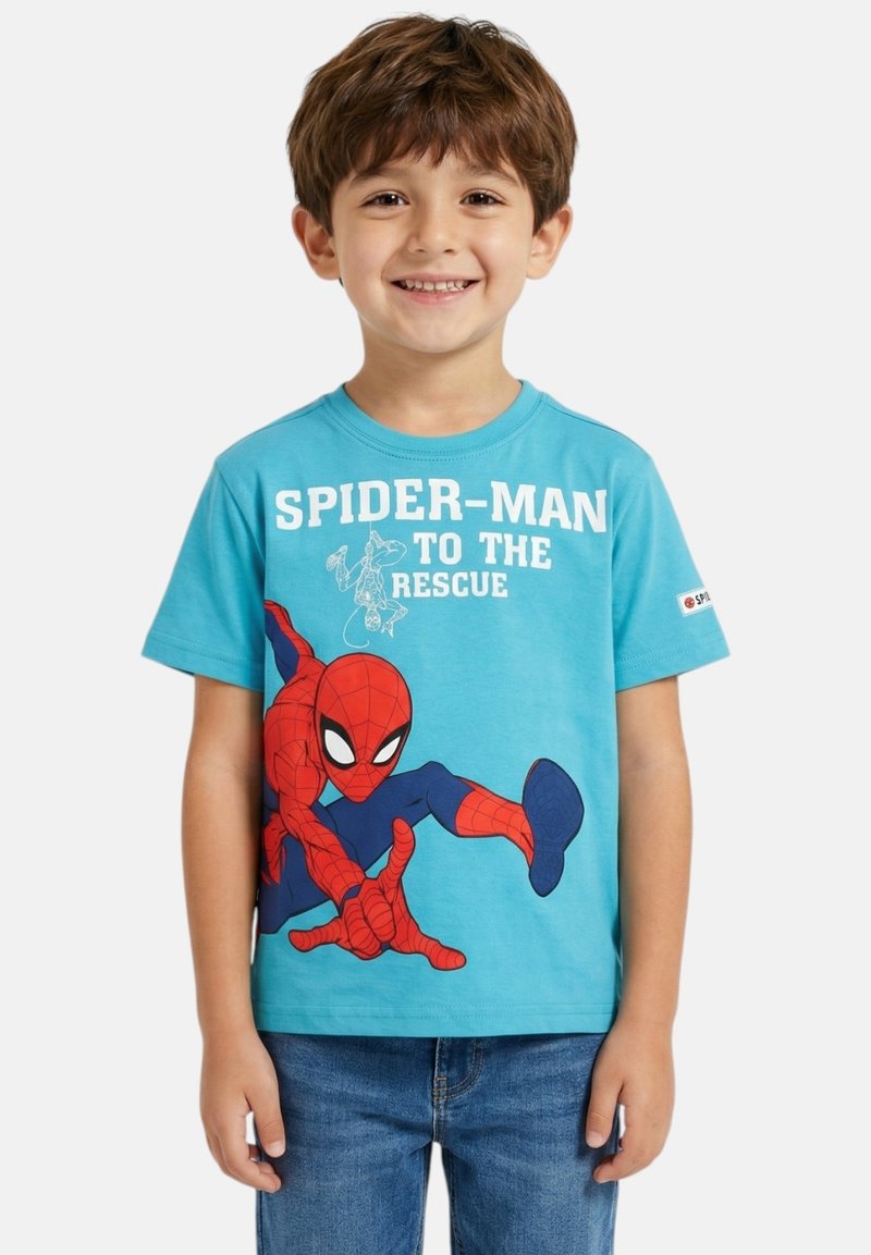Garçon souriant aux cheveux bruns portant un t-shirt bleu Spider-Man et un jean, debout devant un fond clair uni.