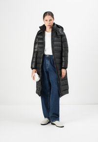 Vrouw in een lange zwarte pufferjas over een witte blouse en blauwe wijde jeans, met een kleine witte tas, staand voor een witte achtergrond.