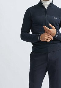 Jersey de color marino con paneles superiores texturizados, cuello con media cremallera y puños acanalados. Presenta el logo de la marca en el pecho y detalles en blanco en el dobladillo.