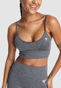 Szary sportowy crop top z teksturowanej tkaniny, z cienkimi ramiączkami i logo po lewej stronie. Zaprojektowany z myślą o komforcie i elastyczności.