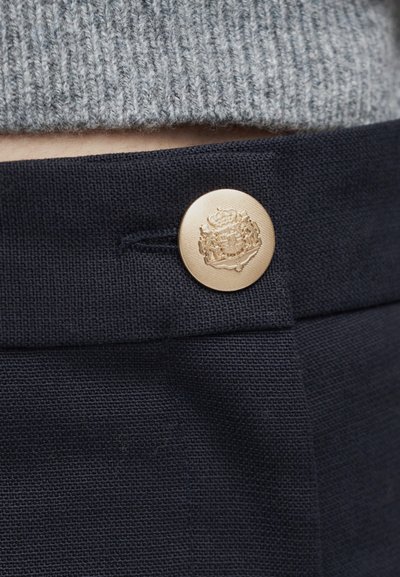 Bouton décoratif doré avec un blason en relief fixant un pantalon en tissu texturé bleu foncé sous un pull gris en tricot.
