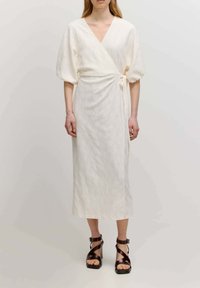 Weißes Wickelkleid mit kurzen Puffärmelchen, floralem Lochmuster und einer Bindegürtel in der Taille. Kombiniert mit braunen, schlichten High-Heel-Sandalen.
