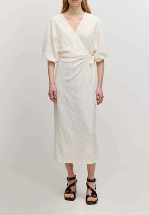 White, wrap-style dress with short puffed sleeves, floral eyelet pattern, and a self-tie waist. Paired with brown strappy high-heel sandals.