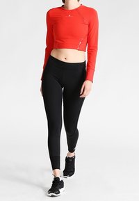 Červený zkrácený dlouhý rukáv sportovní top se švy, doplněný černými těsnými legínami. Modelka má na sobě černé sportovní boty.