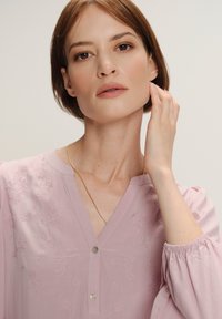 Donna con capelli corti castano ramati, che indossa una blusa ricamata di un rosa chiaro e una delicata collana d'oro, mentre si tocca dolcemente il viso.
